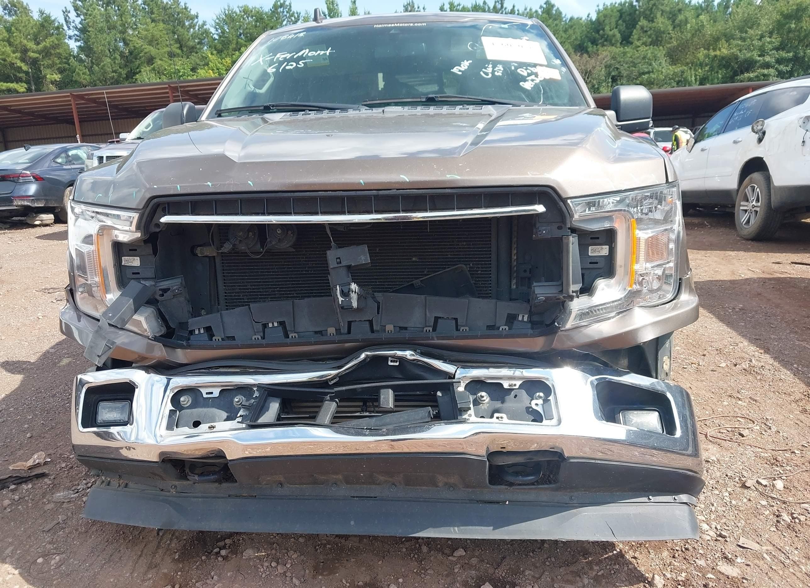 Same 2018 Ford F-150 XLT FX4, head-on angle showing missing radiator support, exposed core support, and detached chrome bumper resting on the ground